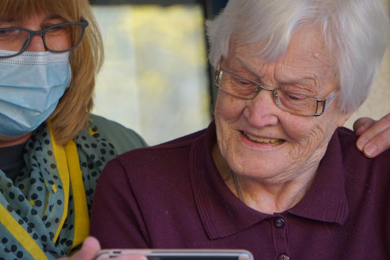 A women wearing a face mask helps an elderly lady read a phone.